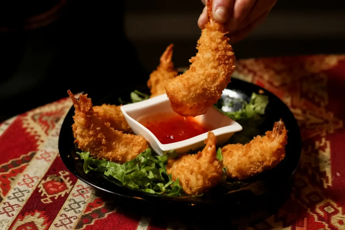 A hand dipping a golden fried shrimp into a dish of vibrant Bang Bang Sauce.