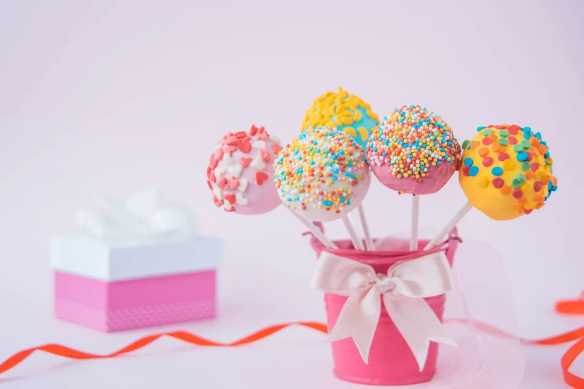 Colorful cake pops adorned with sprinkles, displayed in a pink bucket with a bow, next to a gift box.