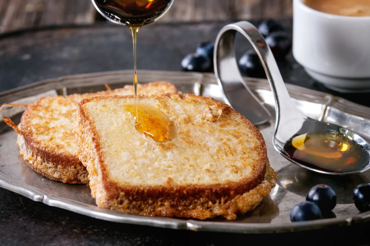 Golden crispy French toast on a vintage silver tray, drizzled with rich maple syrup, accompanied by fresh blueberries and a cup of coffee in the background.