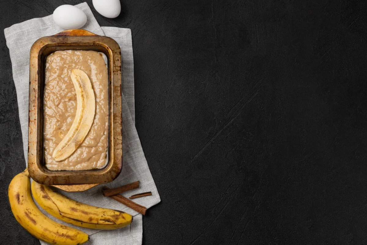 Unbaked banana bread batter in a loaf pan, with ripe bananas and cinnamon sticks on a dark surface.