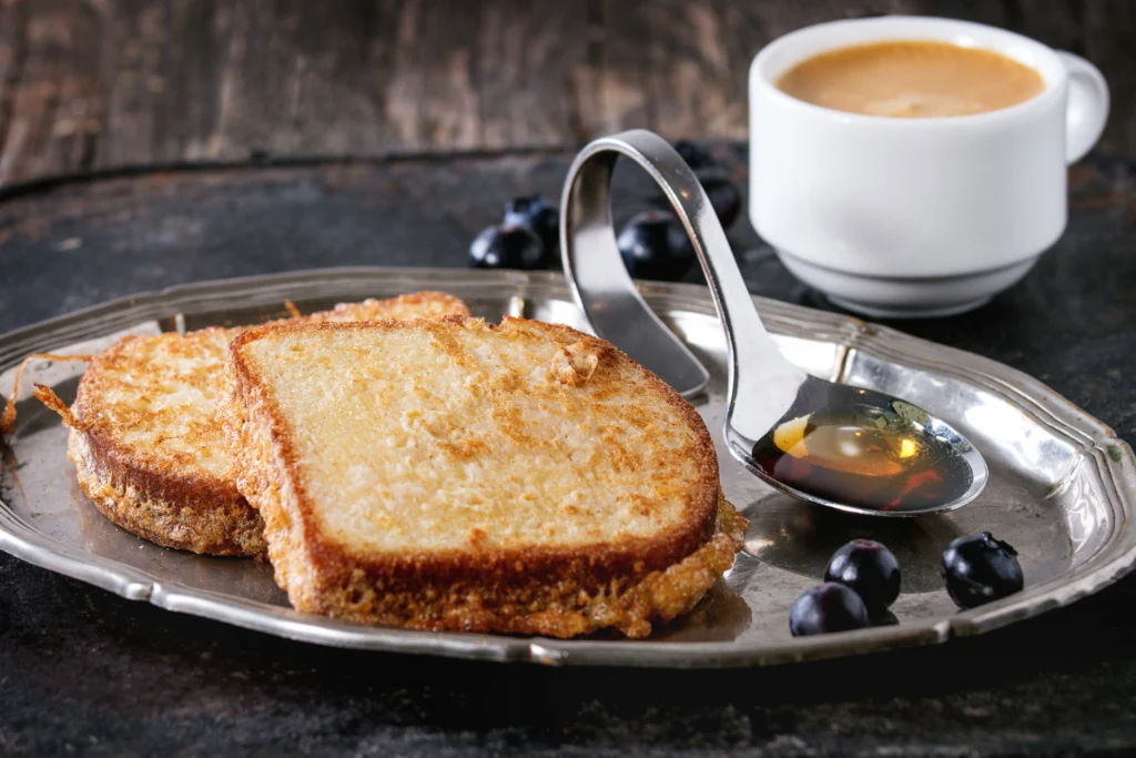 Golden-brown French toast on a vintage silver platter with blueberries and a cup of coffee in the background.