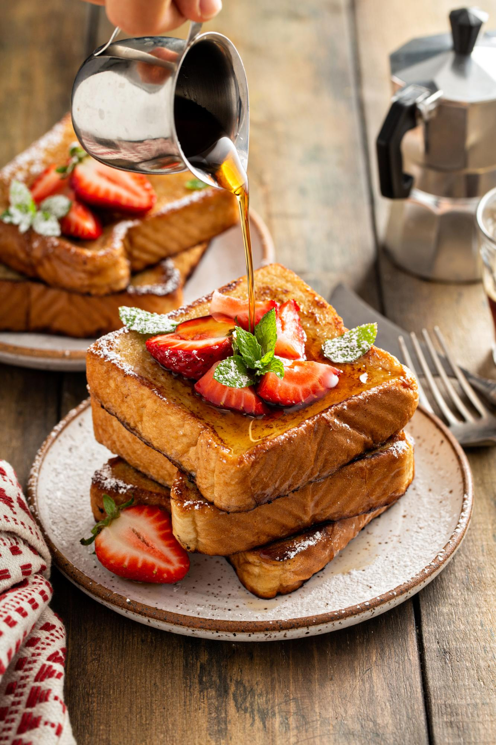 A drizzle of syrup cascading over thick, fluffy French toast adorned with sliced strawberries and a sprig of mint on a rustic table.