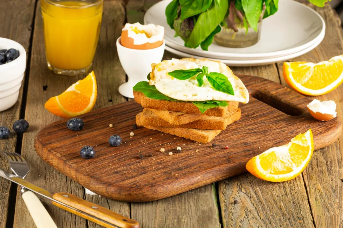 A wholesome breakfast setup featuring toast topped with fried egg and basil, accompanied by fresh blueberries, orange slices, and a glass of orange juice.