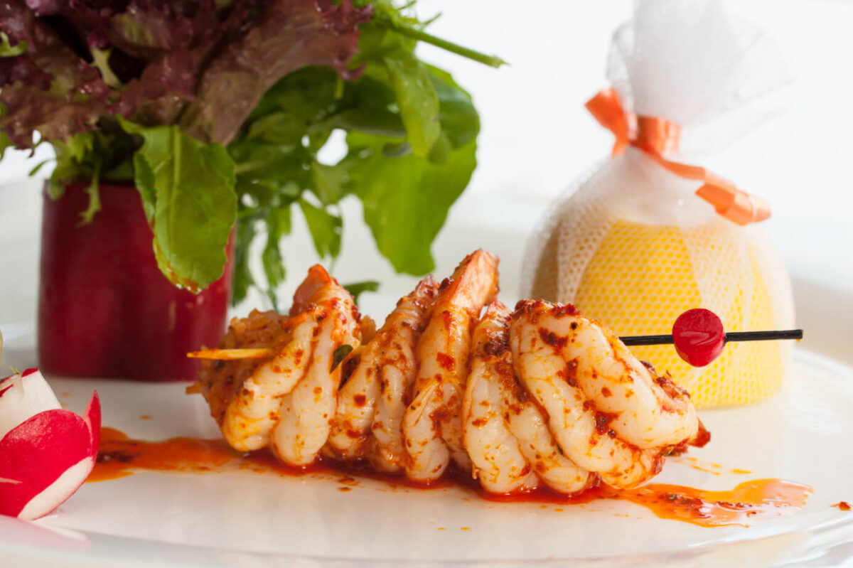 ALT Text: A skewer of succulent Bang Bang Shrimp with spicy sauce, accompanied by fresh greens and a lemon wrapped in mesh, on a white plate.