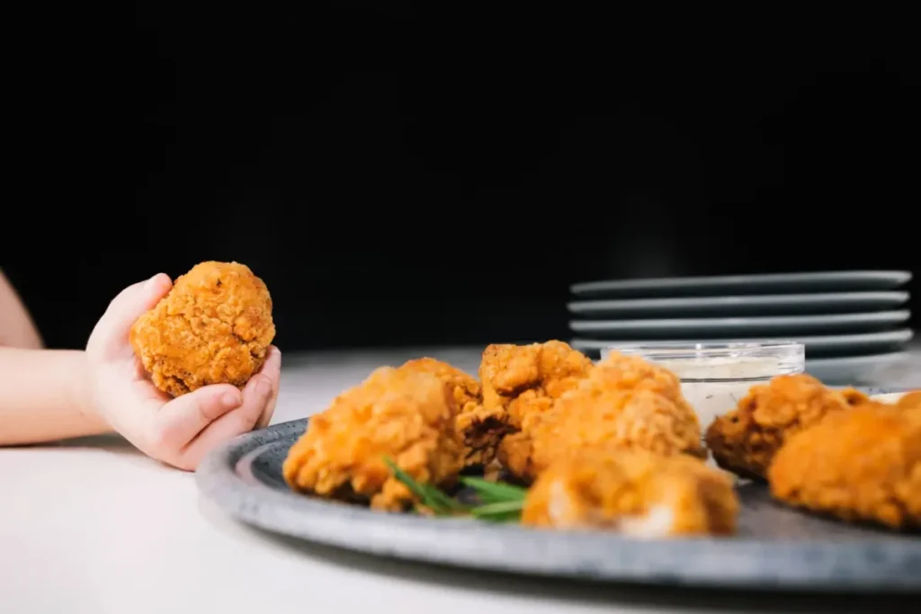 A child's hand holding a piece of crispy fried chicken with more pieces on a plate in the background.