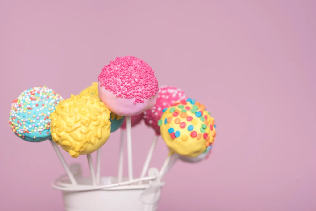 Colorful cake pops with various sprinkles on a pink background.