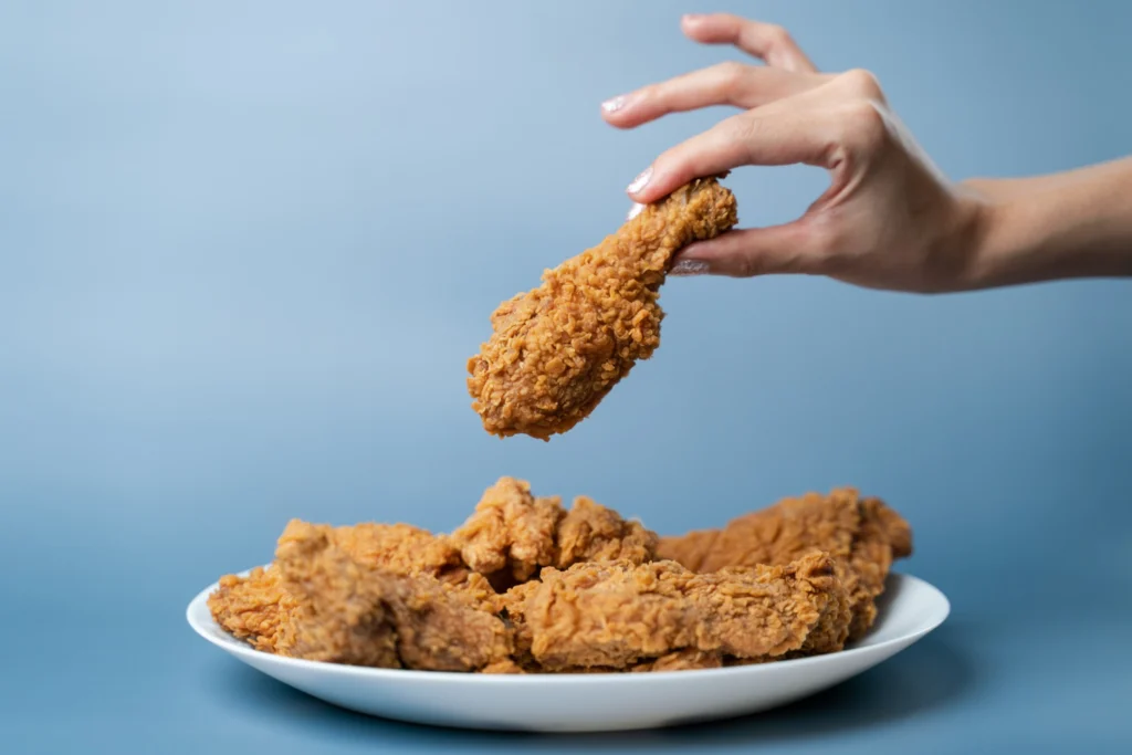 A hand picking up a piece of crispy fried chicken from a white plate against a blue background.