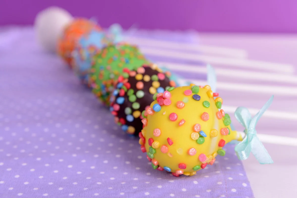 Colorful cake pops with sprinkles and a bow on a polka dot surface.