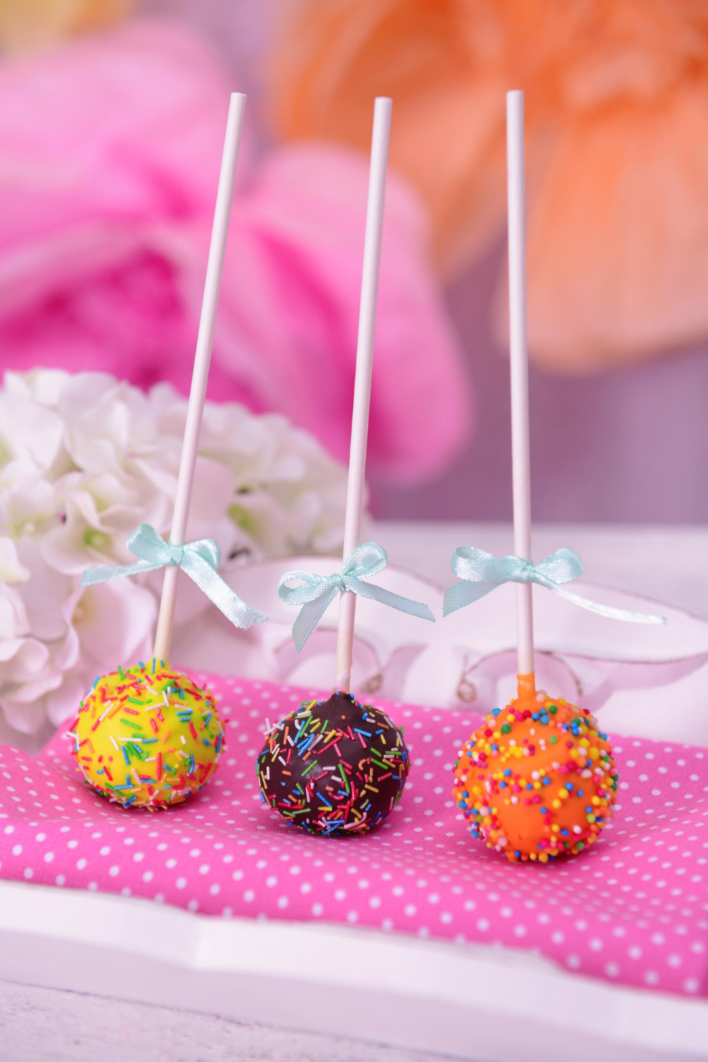 Vibrant cake pops with blue ribbons and assorted sprinkles on a polka-dotted pink surface.