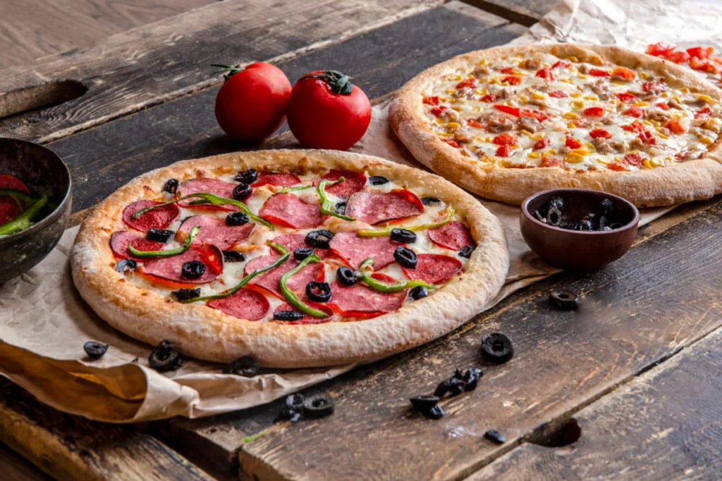 Two freshly baked pizzas with pepperoni, bell peppers, and olives on a rustic wooden table, with tomatoes and olives on the side.