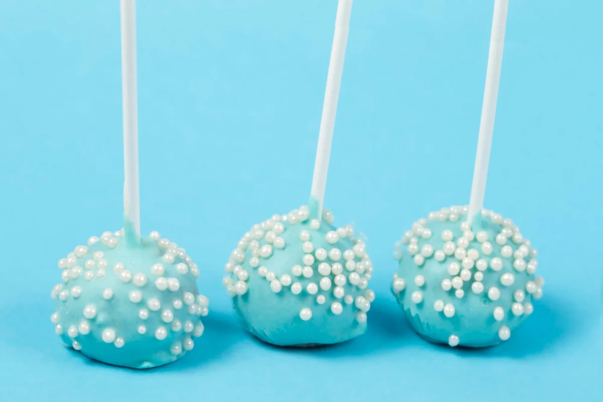 Three cake pops with a pastel blue chocolate coating and white pearl sprinkles against a light blue background.