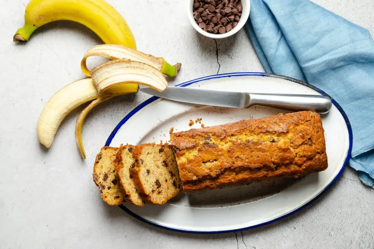 Sliced banana bread with chocolate chips on a white and blue rimmed plate, accompanied by a ripe banana and chocolate chips in a bowl.