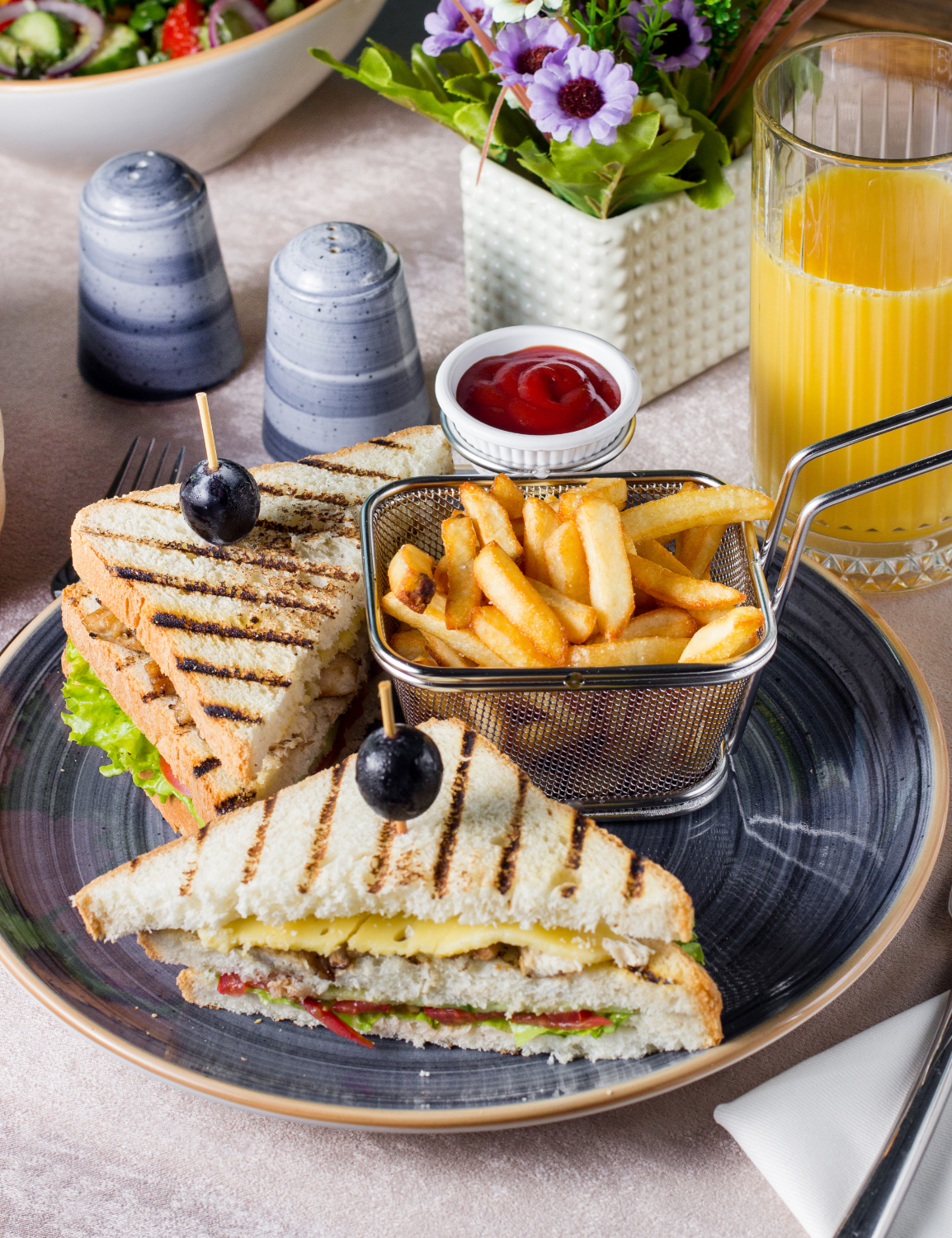 A grilled French toast sandwich with cheese and vegetables, served with fries and orange juice on a dining table.