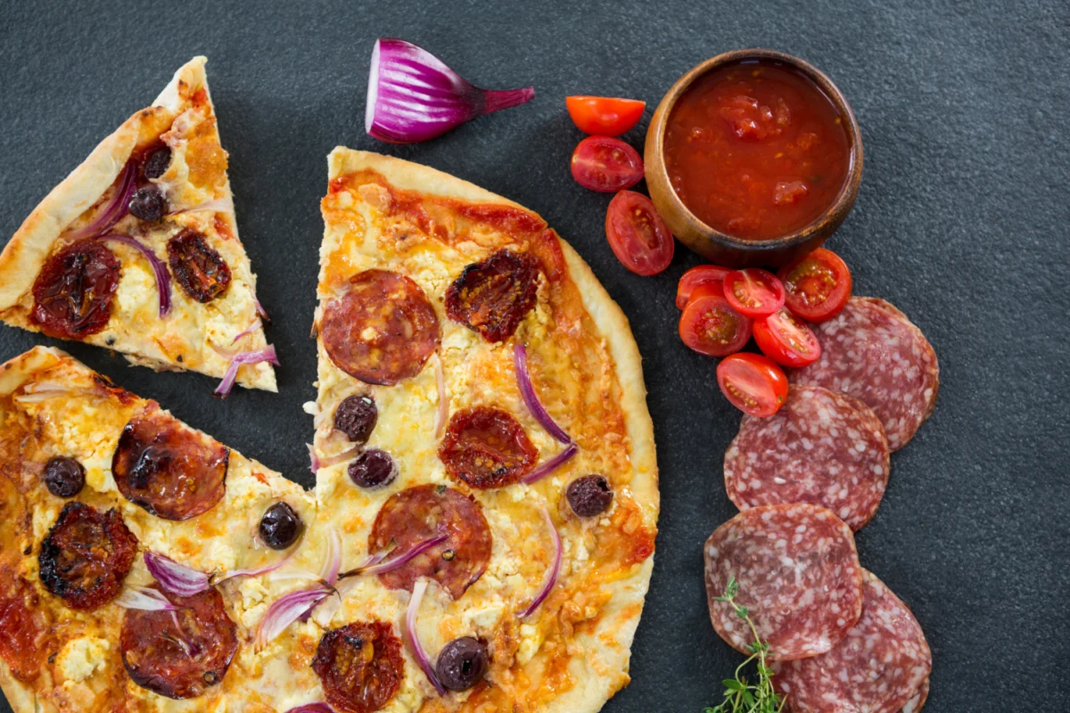 Gourmet pizza slice with pepperoni, onions, and olives next to sliced Sopressata and cherry tomatoes, exploring if they are the same.