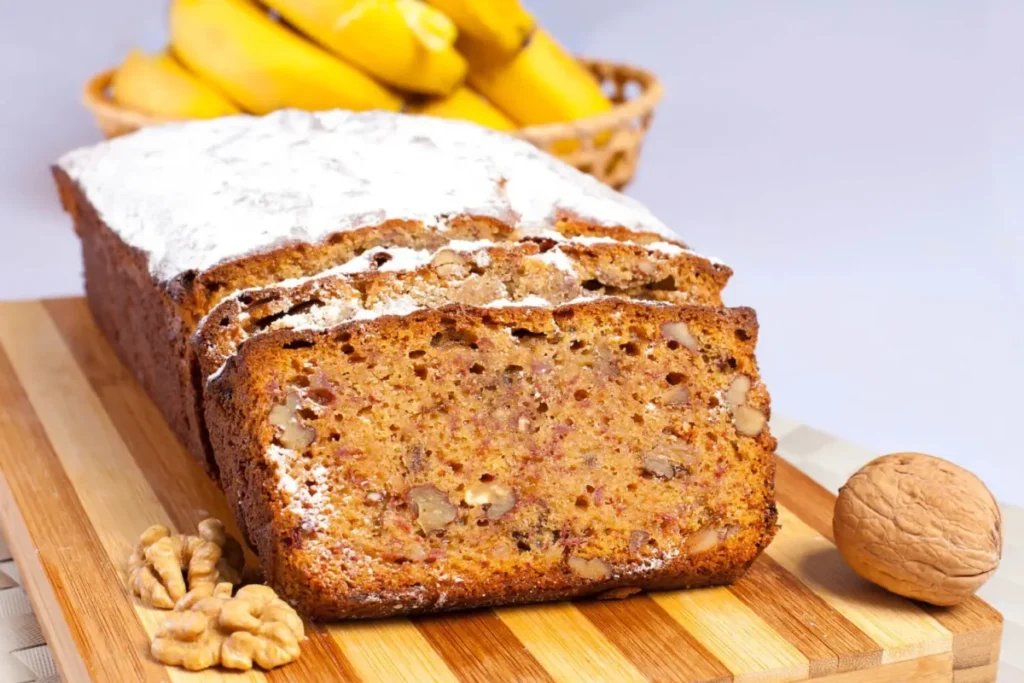 Freshly baked banana bread with walnuts, dusted with powdered sugar, on a wooden cutting board with bananas in the background.