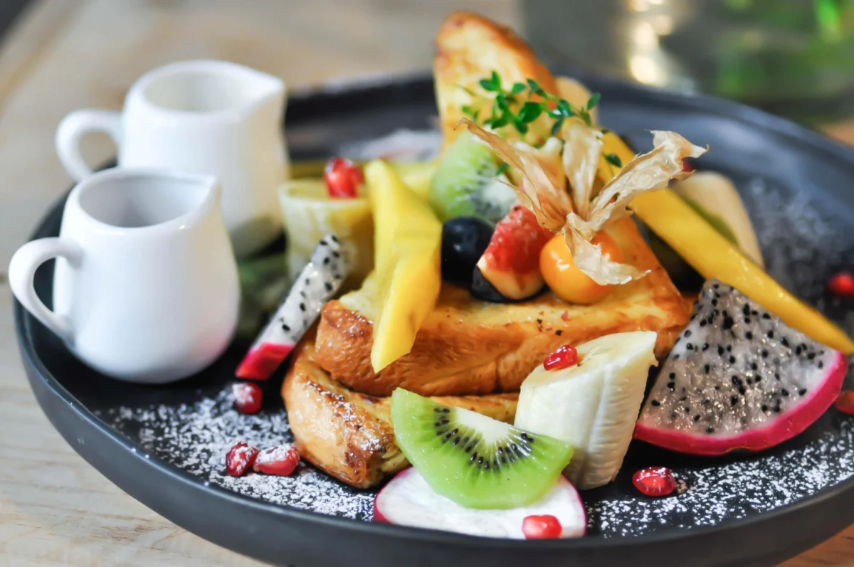 Artfully plated French toast garnished with an array of tropical fruits, sprinkled with powdered sugar, served with syrup and cream on a modern black plate.