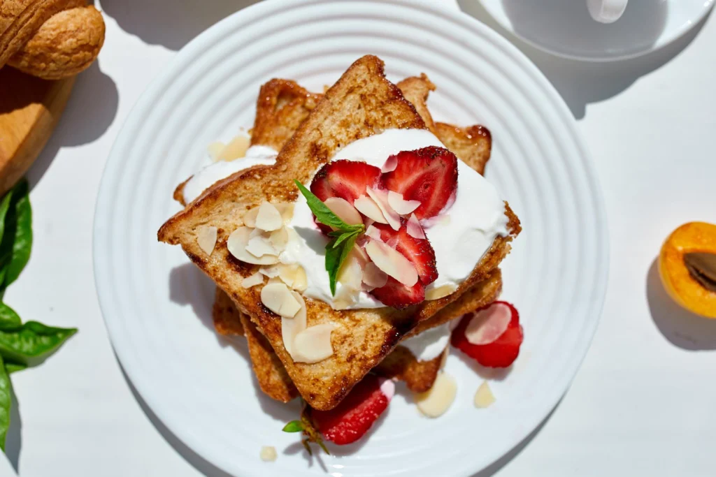 Two slices of golden-brown French toast stacked on a white plate, topped with whipped cream, fresh strawberries, and almond flakes, with a cup and a half apricot in the background.